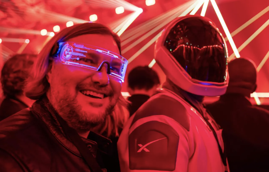A smiling man wearing illuminated glasses next to a person in a SpaceX spacesuit at a red-lit event